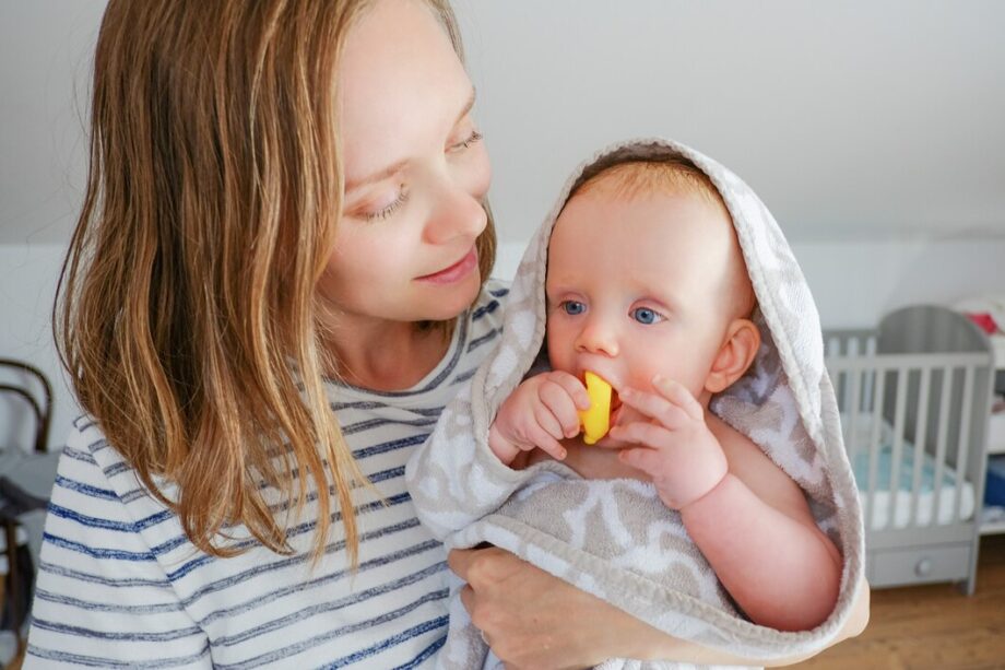 positive-mom-holding-sweet-dry-baby-wearing-hooded-towel-after-shower-biting-rubber-bathing-toy-front-view-child-care-bathing